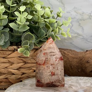May include: A clear quartz crystal point with red inclusions. The crystal is polished and has a pointed tip. It is sitting on a white agate slice.