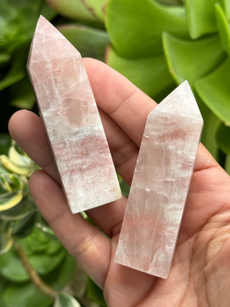 May include: Two white and pink crystal points, also known as selenite wands, are held in a hand. The wands are smooth and have a translucent appearance.