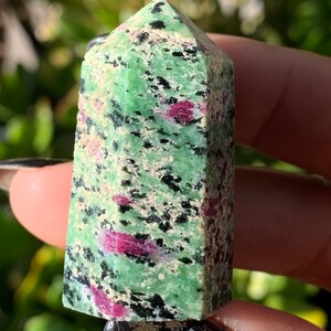 Peut inclure: Une tour de cristal de rubis zoisite verte et rose. Le cristal a une base rectangulaire et un sommet pointu. La couleur verte est parsem&eacute;e de taches noires et roses. Le cristal est tenu dans une main.
