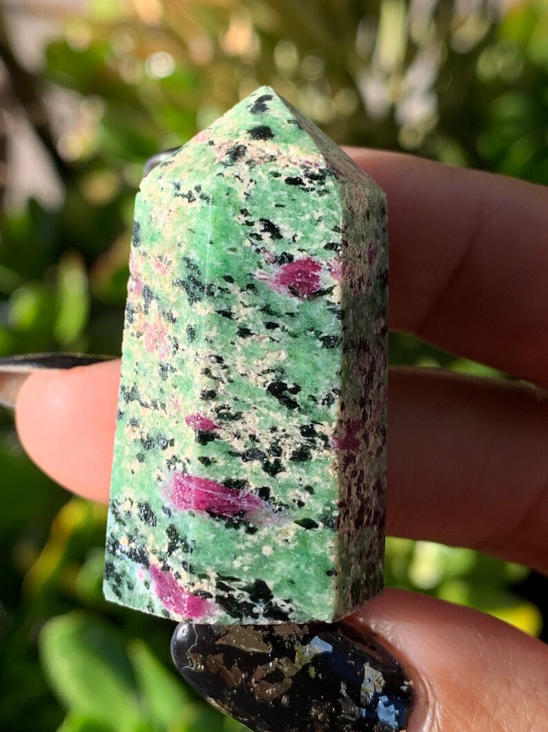 Peut inclure: Une tour de cristal de rubis zoisite verte et rose. Le cristal a une base rectangulaire et un sommet pointu. La couleur verte est parsem&eacute;e de taches noires et roses. Le cristal est tenu dans une main.