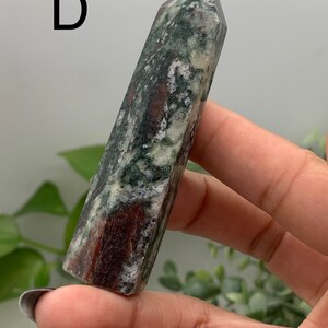 Peut inclure: Une pointe de cristal polie, de forme rectangulaire avec une base plate et un sommet pointu. La pierre pr&eacute;sente un m&eacute;lange de couleurs vertes, marron et blanches. La lettre "D" est visible en haut &agrave; gauche. Le cristal est tenu dans une main.