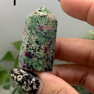 Peut inclure: Une tour en cristal de rubis zoisite, de couleur verte et rouge. La surface du cristal est polie et lisse, avec des inclusions noires. La lettre "F" est visible en arri&egrave;re-plan.