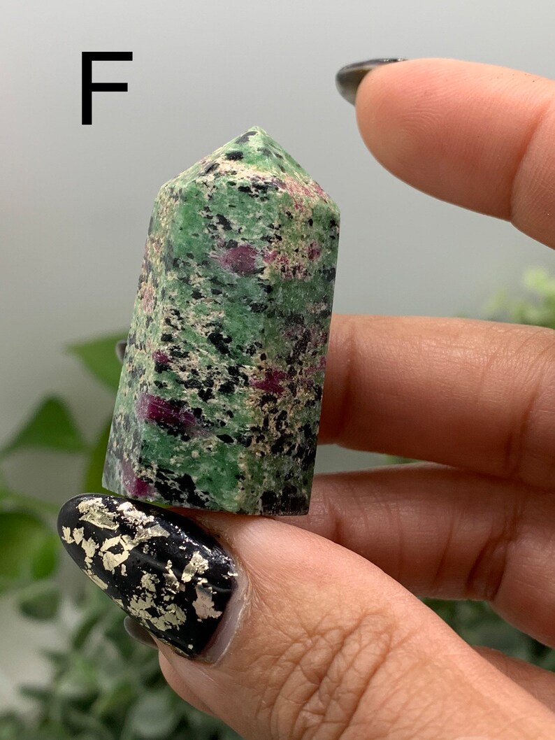 Peut inclure: Une tour en cristal de rubis zoisite, de couleur verte et rouge. La surface du cristal est polie et lisse, avec des inclusions noires. La lettre "F" est visible en arri&egrave;re-plan.