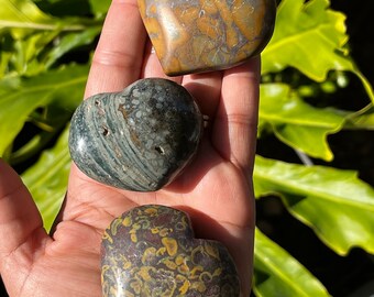 May include: Three heart-shaped polished stones with different patterns. The stones are brown, green, and black with yellow and white markings.