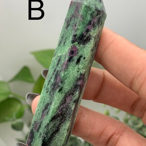 Peut inclure: Une tour en cristal de rubis zoisite polie. La pierre est verte avec des inclusions noires et roses. La lettre "B" est imprim&eacute;e en noir en haut &agrave; gauche de l'image. Le cristal est tenu dans une main.