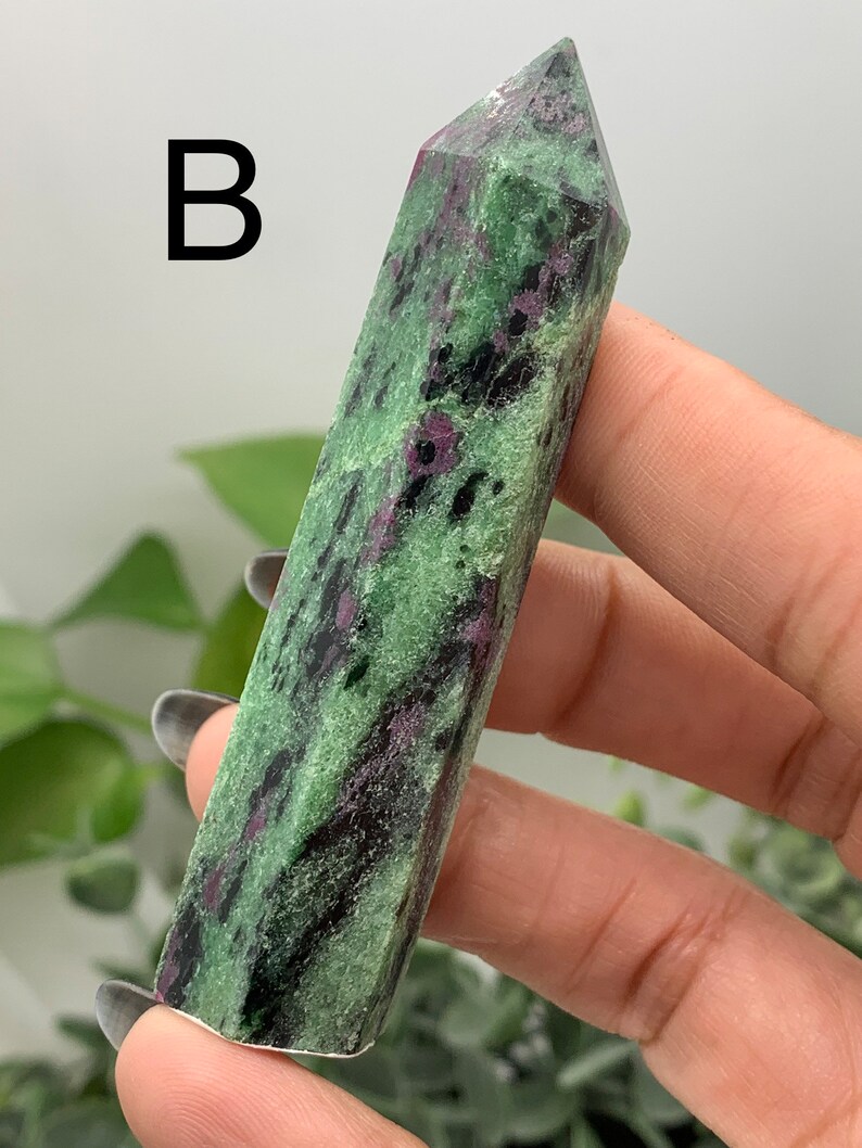 Peut inclure: Une tour en cristal de rubis zoisite polie. La pierre est verte avec des inclusions noires et roses. La lettre "B" est imprim&eacute;e en noir en haut &agrave; gauche de l'image. Le cristal est tenu dans une main.