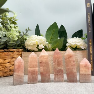 May include: Seven pink rose quartz crystal points arranged in a row on a gray surface. The crystals are all different sizes and shapes, but they are all polished and have a smooth, shiny finish.
