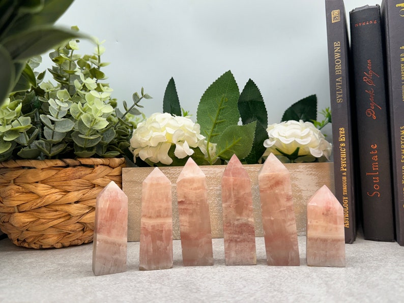 May include: Seven pink rose quartz crystal points arranged in a row on a gray surface. The crystals are all different sizes and shapes, but they are all polished and have a smooth, shiny finish.