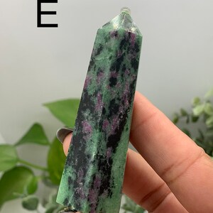 Peut inclure: Une tour de cristal de zo&iuml;site verte et noire avec des &eacute;clats roses. Le cristal est tenu dans une main et a une surface polie et lisse. La lettre "E" est dans le coin sup&eacute;rieur gauche.