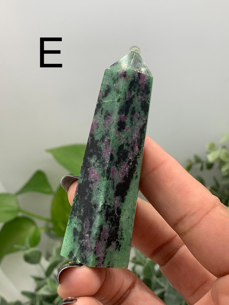 Peut inclure: Une tour de cristal de zo&iuml;site verte et noire avec des &eacute;clats roses. Le cristal est tenu dans une main et a une surface polie et lisse. La lettre "E" est dans le coin sup&eacute;rieur gauche.