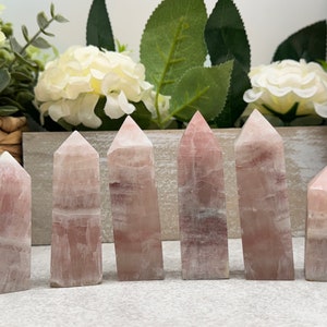 May include: Seven pink and white crystal points, also known as wands, are arranged in a row on a light gray surface. The points are all different sizes and shapes, but they are all made of the same type of crystal.