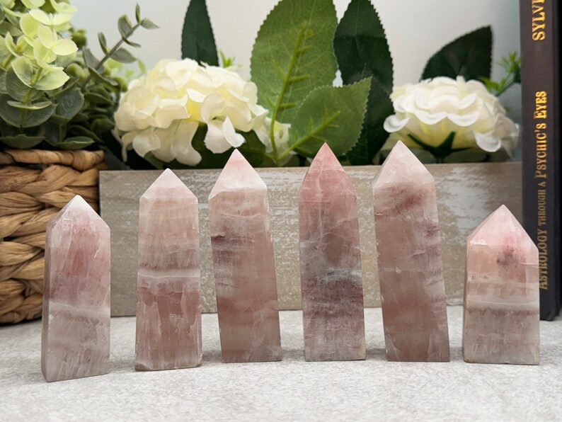 May include: Seven pink and white crystal points, also known as wands, are arranged in a row on a light gray surface. The points are all different sizes and shapes, but they are all made of the same type of crystal.