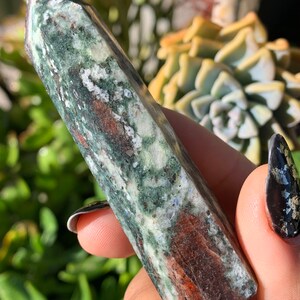 Peut inclure: Un ob&eacute;lisque en cristal vert, marron et blanc avec un sommet pointu. Le cristal a un aspect naturel marbr&eacute; avec des variations de couleur et de texture. L'ob&eacute;lisque est tenu dans une main, avec un arri&egrave;re-plan flou de plantes vertes.