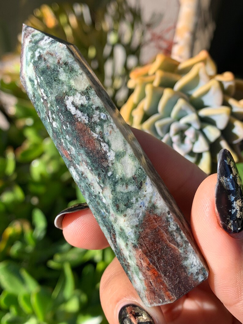 Peut inclure: Un ob&eacute;lisque en cristal vert, marron et blanc avec un sommet pointu. Le cristal a un aspect naturel marbr&eacute; avec des variations de couleur et de texture. L'ob&eacute;lisque est tenu dans une main, avec un arri&egrave;re-plan flou de plantes vertes.