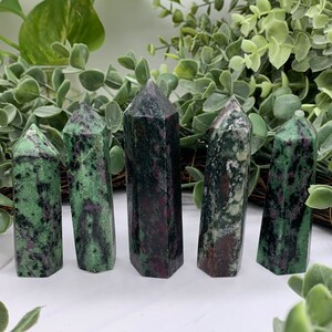 Peut inclure: Cinq tours de cristal de zoisite polies. Les cristaux sont un m&eacute;lange de vert et de noir avec des inclusions rouges. Les tours sont dress&eacute;es, avec une vari&eacute;t&eacute; de formes et de tailles. Le fond pr&eacute;sente un feuillage vert.