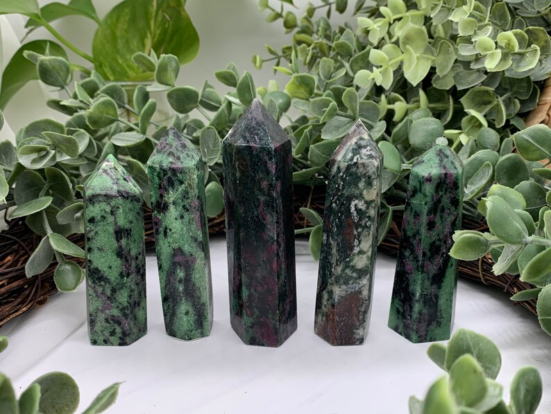 Peut inclure: Cinq tours de cristal de zoisite polies. Les cristaux sont un m&eacute;lange de vert et de noir avec des inclusions rouges. Les tours sont dress&eacute;es, avec une vari&eacute;t&eacute; de formes et de tailles. Le fond pr&eacute;sente un feuillage vert.