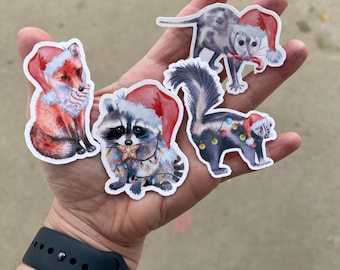 May include: A collection of five animal stickers, including a fox, raccoon, opossum, and skunk, all wearing Santa hats. The raccoon is adorned with string lights. The stickers are colorful and whimsical, perfect for holiday decorations.