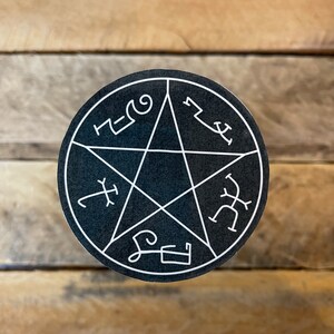 May include: A round, black sticker featuring a white pentagram design with stylized symbols around the edges. The sticker is centered on a wooden surface, creating a contrast between the dark sticker and the light wood.