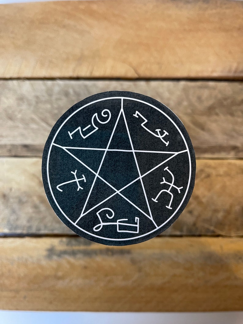 May include: A round, black sticker featuring a white pentagram design with stylized symbols around the edges. The sticker is centered on a wooden surface, creating a contrast between the dark sticker and the light wood.
