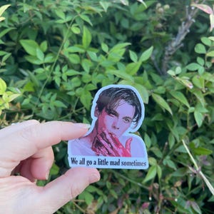 May include: A sticker featuring a portrait of a man with dark hair and a red stain on his hand. The sticker has the text "We all go a little mad sometimes" printed below the image.