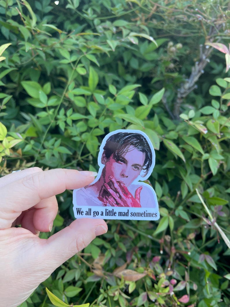 May include: A sticker featuring a portrait of a man with dark hair and a red stain on his hand. The sticker has the text "We all go a little mad sometimes" printed below the image.