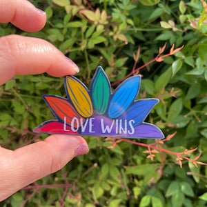 May include: A rainbow-colored sticker with the words "LOVE WINS" in white lettering. The sticker is shaped like a flower with multiple petals.