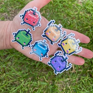 May include: Six pixelated character stickers with a glittery finish. The characters are in various colors, including red, green, orange, yellow, blue, and purple. Each character has a unique expression and is shaped like a small, rounded creature.