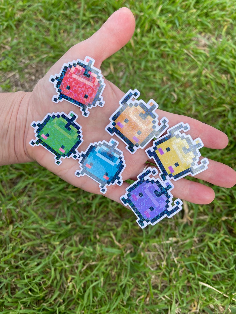 May include: Six pixelated character stickers with a glittery finish. The characters are in various colours, including red, green, orange, yellow, blue, and purple. Each character has a unique expression and is shaped like a small, rounded creature.