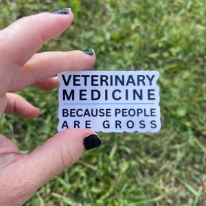 May include: A white sticker with black text that reads "VETERINARY MEDICINE BECAUSE PEOPLE ARE GROSS." The sticker is rectangular with a wavy border and is held in front of a grassy background.