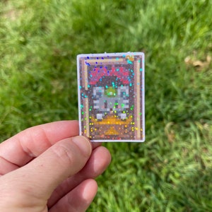 May include: A holographic sticker featuring a pixelated image of a chest with a golden lid. The chest is surrounded by a brown frame with a red and white border.