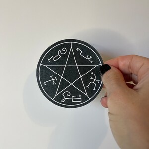 May include: A black, circular sticker featuring a white pentagram design with various symbols. The sticker is held by a hand with black nail polish against a white background.