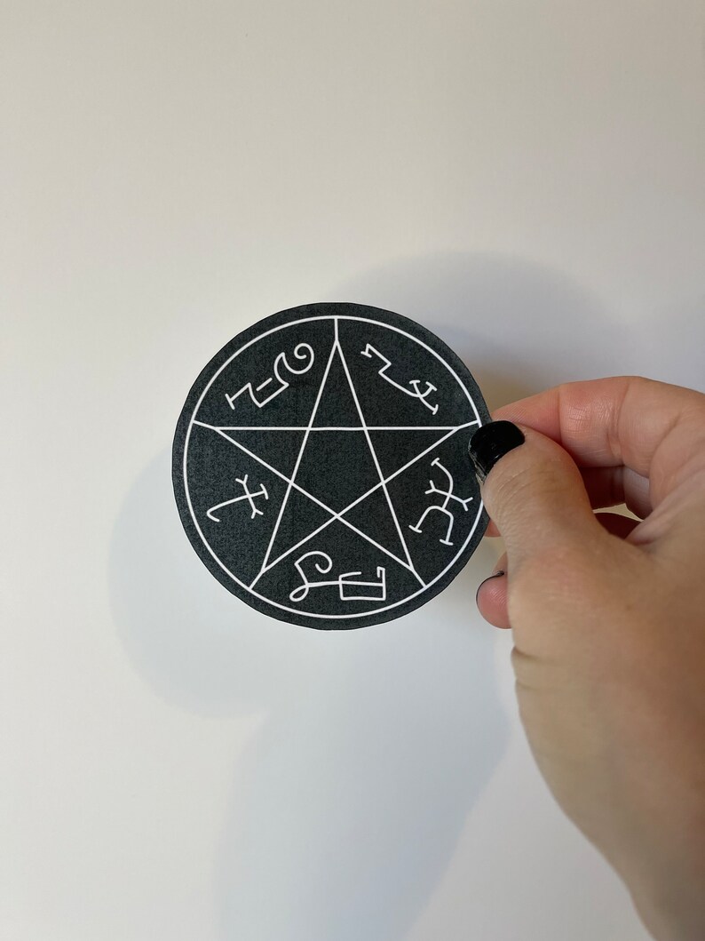 May include: A black, circular sticker featuring a white pentagram design with various symbols. The sticker is held by a hand with black nail polish against a white background.