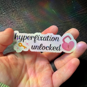 May include: A holographic sticker with the text "hyperfixation unlocked" in black font. The design includes a small orange key icon with sparkles on the left and a pink heart-shaped padlock icon on the right, all set against a shimmering background.