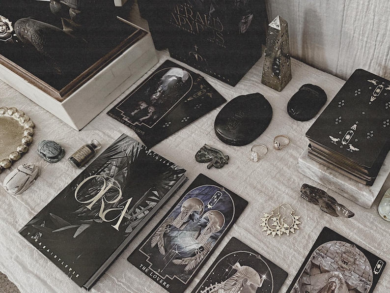 K&ouml;nnte beinhalten: Eine Ansammlung mystischer Gegenst&auml;nde, darunter ein Buch mit dem Titel "ORA", Tarotkarten und dekorative Steine, angeordnet auf einer neutralen Oberfl&auml;che. Die Artikel haben eine dunkle, gotische &Auml;sthetik mit Gold- und Silberakzenten.