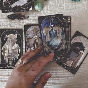 K&ouml;nnte beinhalten: Ein Satz Tarotkarten mit &auml;gyptisch inspirierten Illustrationen. Die Karten zeigen Bilder von Figuren, Symbolen und Text, darunter "The Lovers" und "The High Priestess". Die Farbpalette umfasst Schwarz, Gold und Blau.