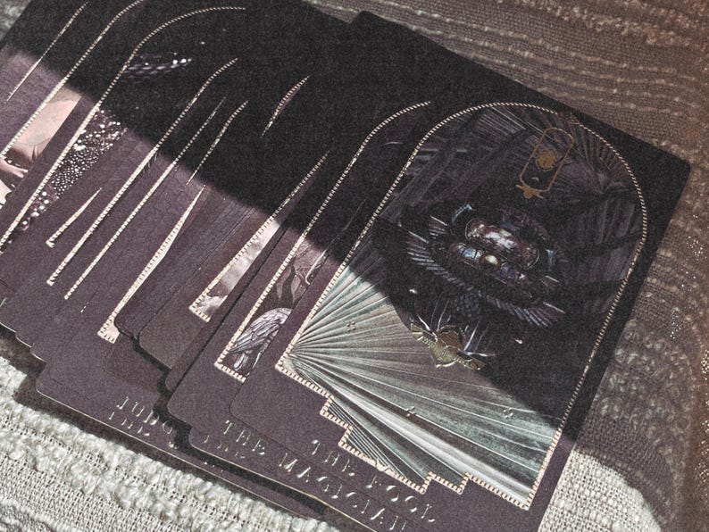 K&ouml;nnte beinhalten: Ein Tarotkartenspiel mit dunkelvioletten R&uuml;ckseiten und silbernen R&auml;ndern. Die Karten zeigen detaillierte Illustrationen in Schwarz-, Grau- und Silbert&ouml;nen. Die Karten sind aufgef&auml;chert, mit den Titeln "Der Magier" und "Der Narr" sichtbar.