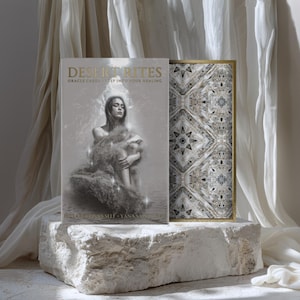 May include: An oracle deck and box set titled "Desiderites: Oracle Cards of Intuitive Healing" featuring a woman illustration. The box has a gold border and a geometric pattern. The items are displayed on a white stone block with draped fabric.