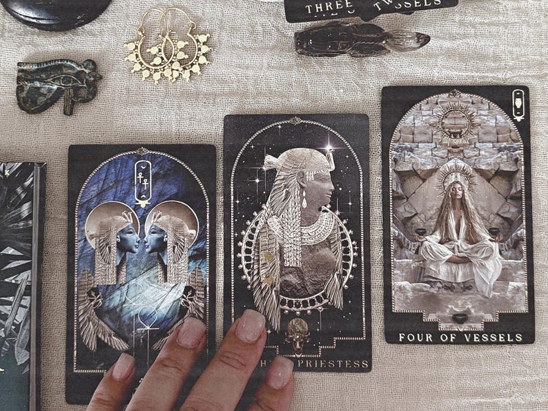 K&ouml;nnte beinhalten: Tarotkarten mit &auml;gyptischen Illustrationen. Die Karten zeigen Bilder von Figuren, Symbolen und die Texte "HIGH PRIESTESS" und "FOUR OF VESSELS". Goldene Ohrringe und andere dekorative Gegenst&auml;nde sind ebenfalls zu sehen.