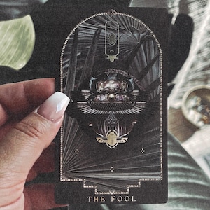 May include: A tarot card with a dark background and gold accents. The card features an illustration of a scarab beetle with wings, set within an arched frame. The text "THE FOOL" is at the bottom. The card is held by a hand with a white manicure.
