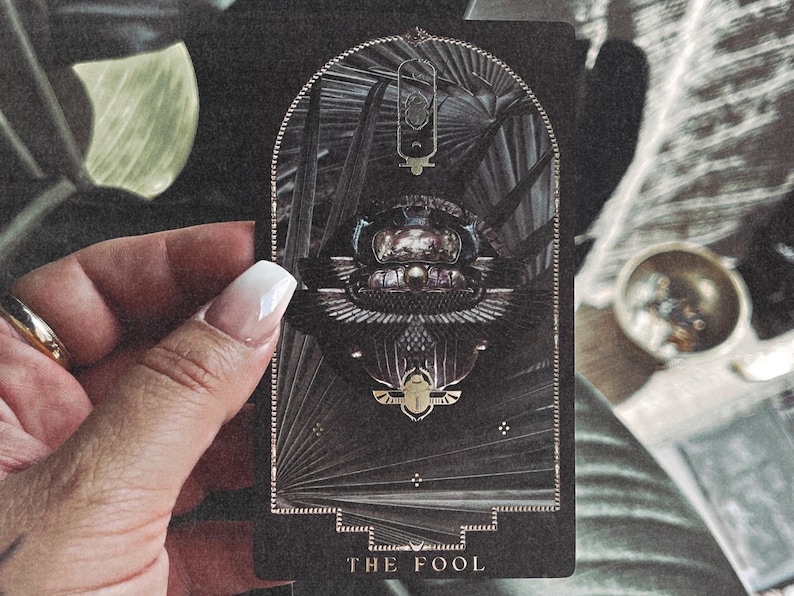 K&ouml;nnte beinhalten: Eine Tarotkarte mit dunklem Hintergrund und goldenen Akzenten. Die Karte zeigt ein verziertes Design mit einem zentralen Bild eines Skarab&auml;us-K&auml;fers und dem Text "THE FOOL" unten. Die Karte wird von einer Hand mit wei&szlig;em Nagellack gehalten.