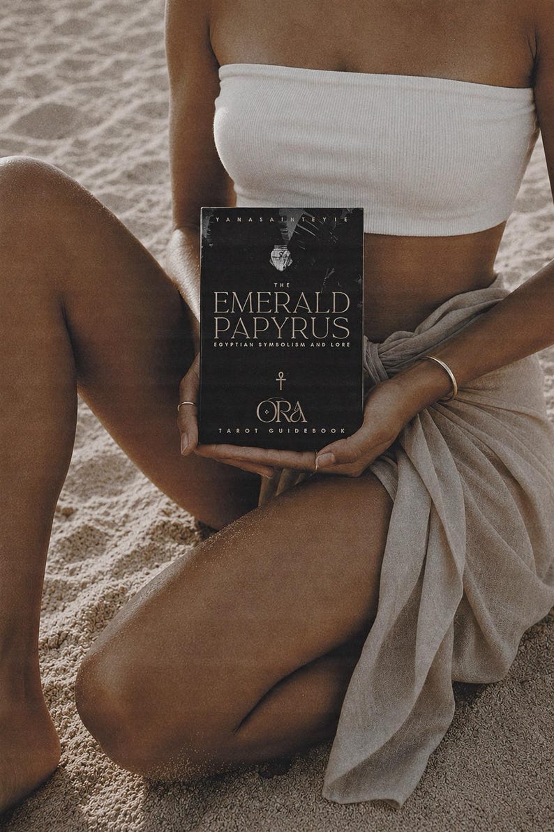 K&ouml;nnte beinhalten: Ein schwarzes Tarot-Handbuch mit dem Titel "The Emerald Papyrus" mit goldener Schrift und &auml;gyptischen Symbolen. Das Buch wird von einer Person gehalten, die ein wei&szlig;es Bandeau-Top tr&auml;gt und auf einer sandigen Oberfl&auml;che sitzt.