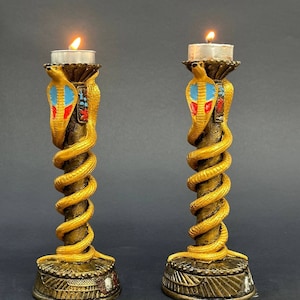 Op de afbeelding: Twee gouden kaarsenstandaards met een spiraalvormig slangendesign. De slangen zijn versierd met rode, blauwe en gouden accenten. De kaarsenstandaards staan op een zwarte achtergrond.