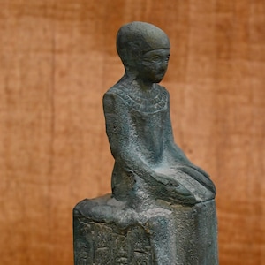 Puede incluir: Una pequeña estatua verde de una figura egipcia sentada con jeroglíficos en la base.