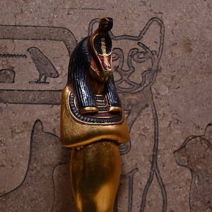 May include: A gold-painted figurine of an Egyptian god with a blue and red headdress. The figurine is standing on a black base.