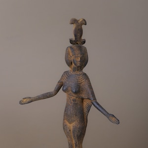 May include: A dark brown, standing figurine of an Egyptian goddess with a headdress. The figurine is made of a textured material and has a detailed design.