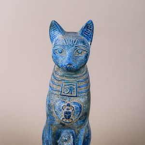 May include: A blue ceramic statue of an Egyptian cat goddess Bastet. The statue is sitting on a base and has hieroglyphics on its body.