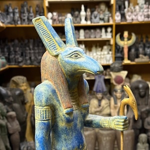 May include: A blue and gold statue of the Egyptian god Anubis, depicted as a jackal-headed human with a staff. The statue is standing on a wooden base.