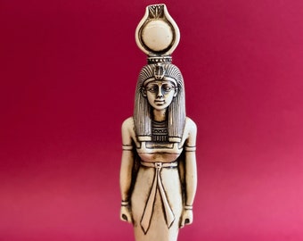 Puede incluir: Figurilla de cerámica blanca de una diosa egipcia con un alto tocado y una larga túnica fluida. La figurilla está de pie sobre una base con jeroglíficos.