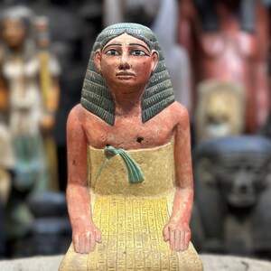 Puede incluir: Una pequeña estatua de una figura egipcia arrodillada con una túnica amarilla y azul, pintada con jeroglíficos. La figura tiene una peluca azul y una cara pintada.