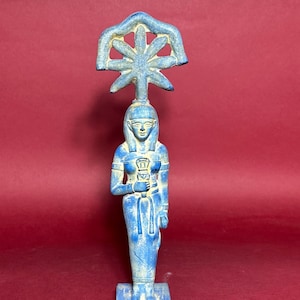 May include: A blue ceramic statue of an Egyptian goddess holding a staff with a sun symbol on top. The statue is standing on a rectangular base with hieroglyphs.